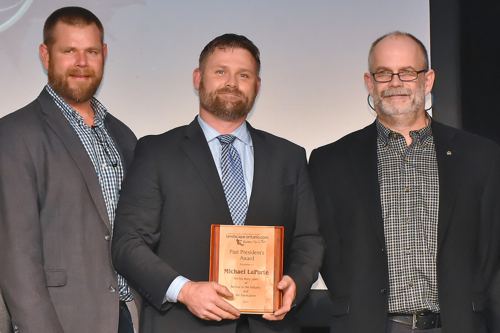 three men on stage. middle one holding an award